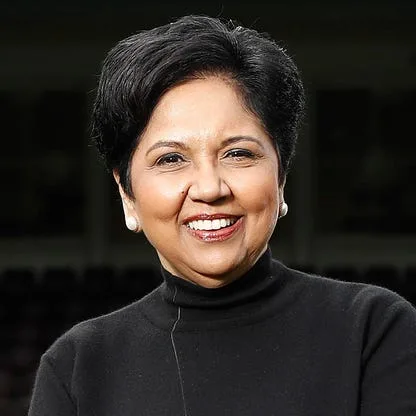 A person with short black hair, wearing a black turtleneck and pearl earrings, is smiling warmly, exuding the charm of a curated customer experience. The background is dark and out of focus.