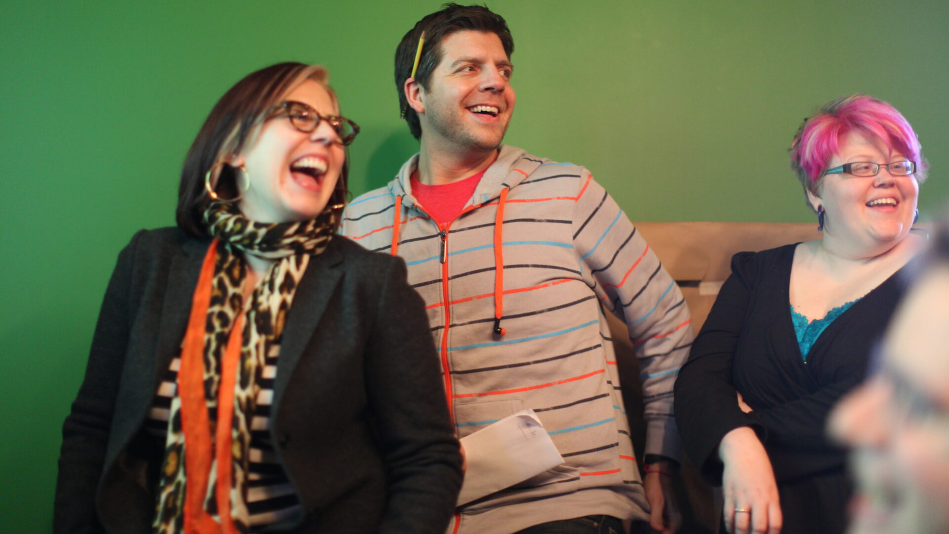Three people are smiling and laughing in front of a vibrant green wall. One sports glasses, a scarf, and an orange shirt; another wears a striped hoodie; the third has pink hair and a black top. Their camaraderie is as seamless as clockwork, capturing a beautifully synchronized moment together.