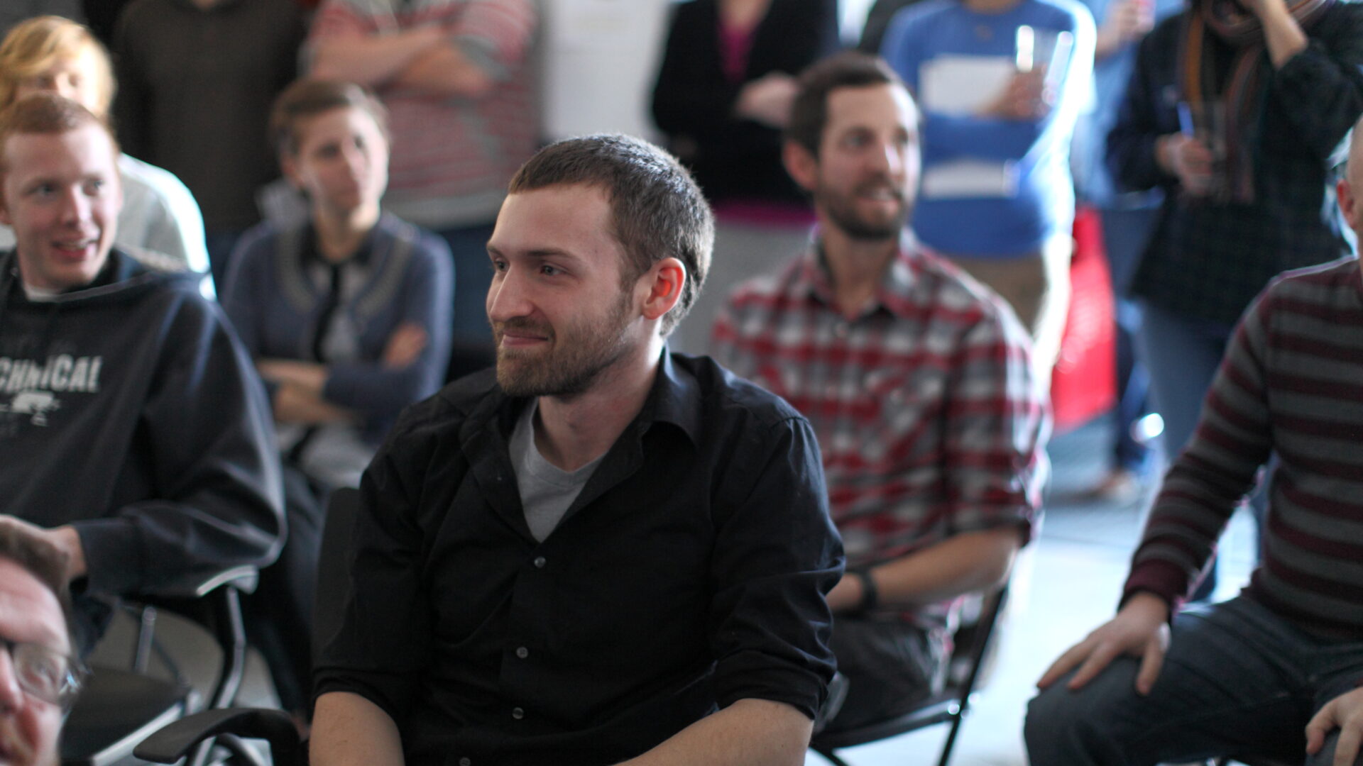 In a casual and relaxed atmosphere, a group of people are seated and standing in a room, engaged and smiling. The focus is on a person in the foreground wearing a black shirt, sitting and looking to the right, perhaps discussing an innovative digital solution.
