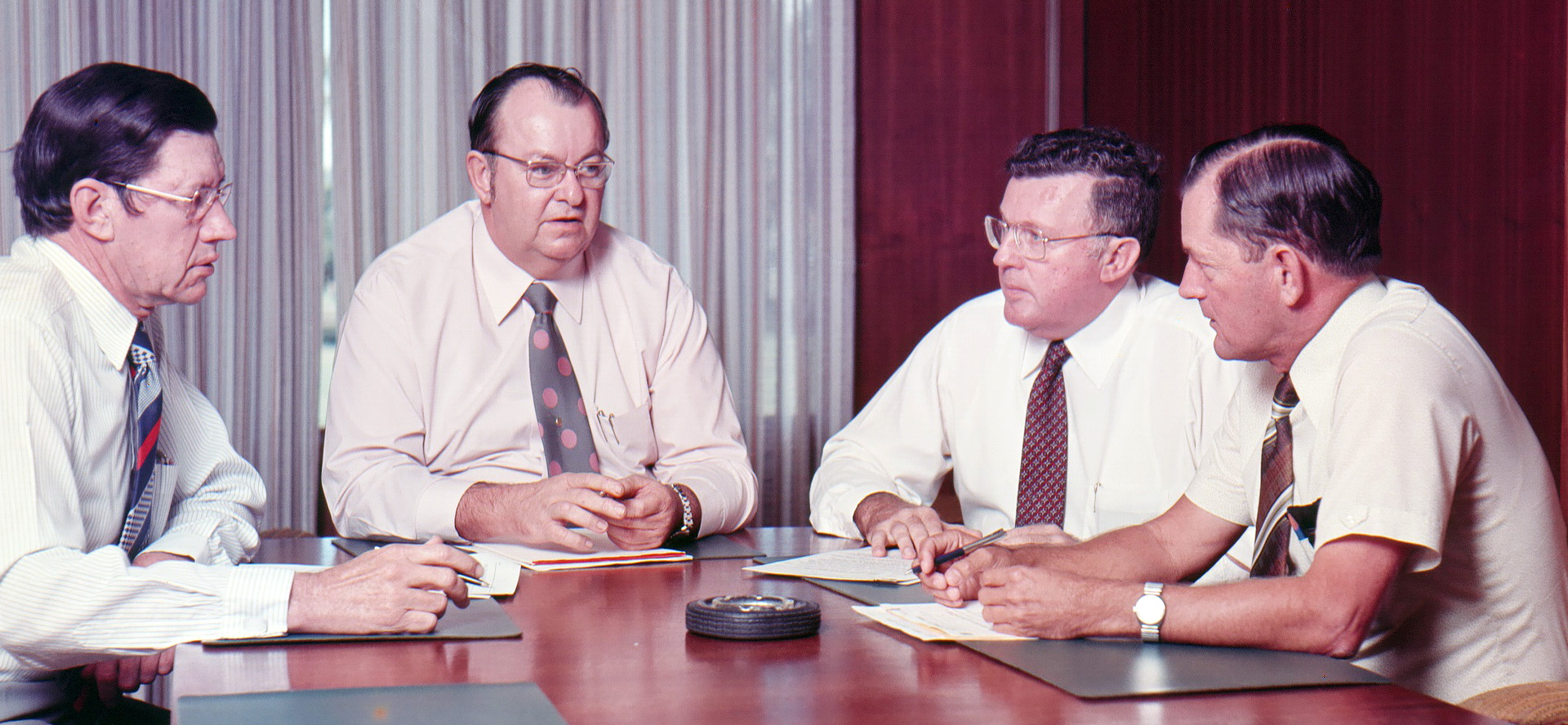 Four men in dress shirts and ties sit around a wooden table engaged in a discussion about user experience. Papers and a small round device are on the table, hinting at custom software strategies. The background features curtains and a wooden panel.