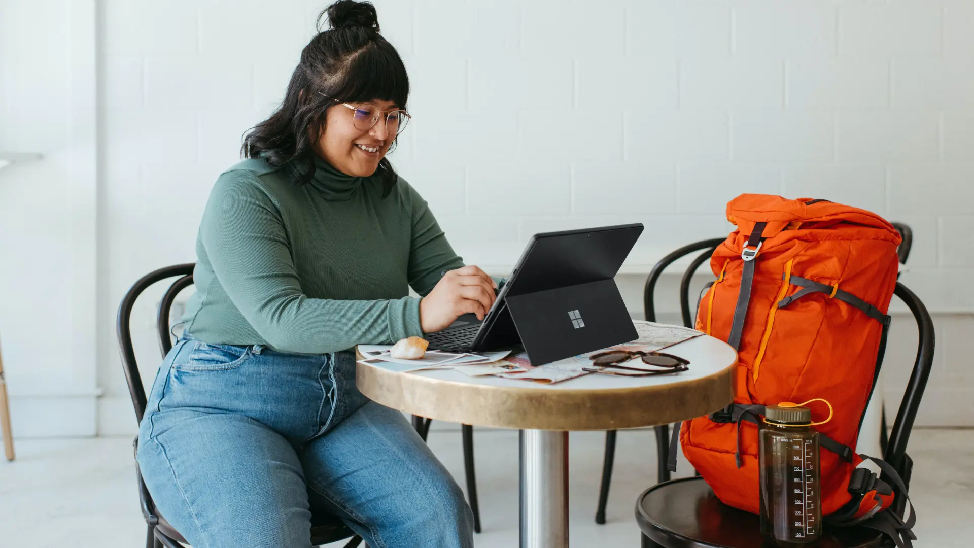 A person with glasses and a green turtleneck is sitting at a table using a tablet, exploring a digital solution. An orange backpack and water bottle sit next to them, enhancing the user experience in this casual indoor space.