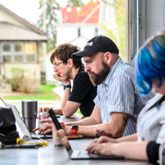 Clockworkers seated at our kitchen table working side by side. The garage door to our Rayvic building is open on a sunny day.