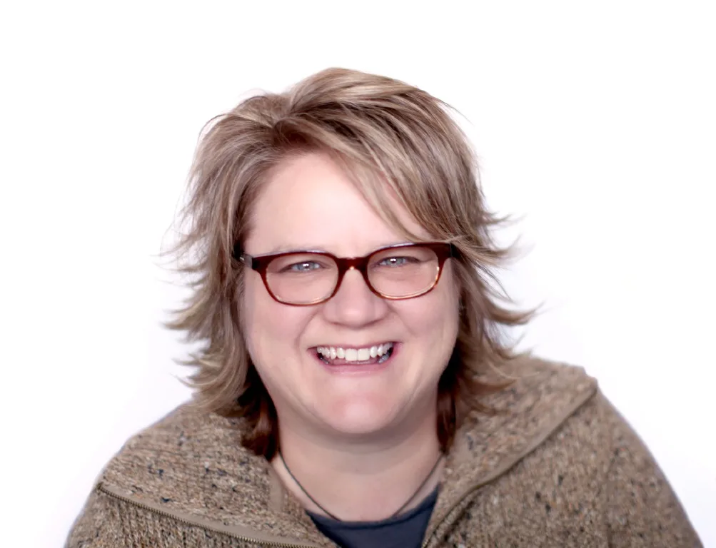 A person with short, tousled hair and glasses smiles at the camera, exuding a clockwork charm. They wear a speckled sweater with a zipper against a plain white background, creating an effortless blend of warmth and style.