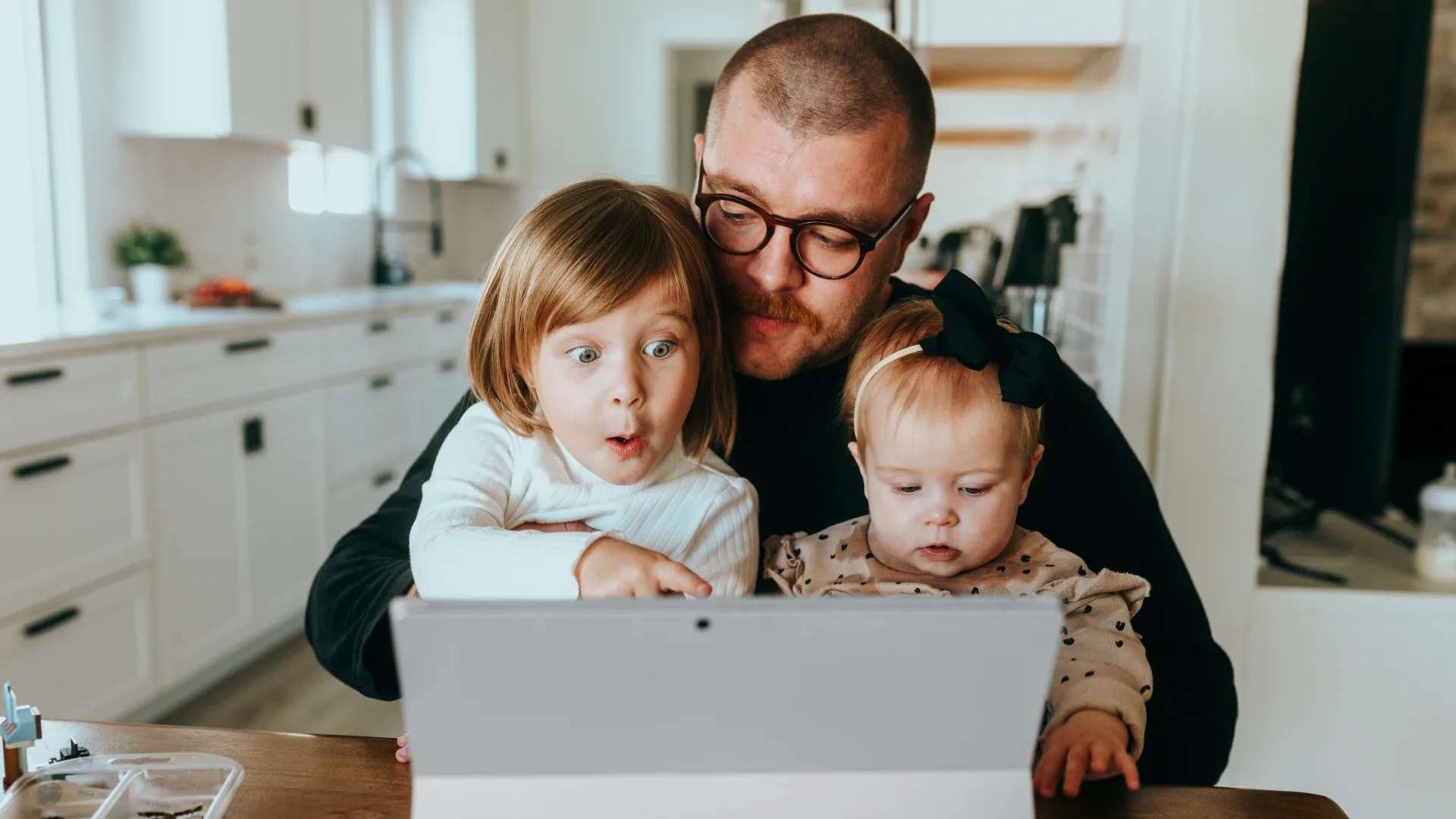 A man with glasses sits at a table with two young children, exploring a custom software on a tablet. The older child appears excited while the toddler remains focused, enhancing their customer experience in the bright kitchen setting.