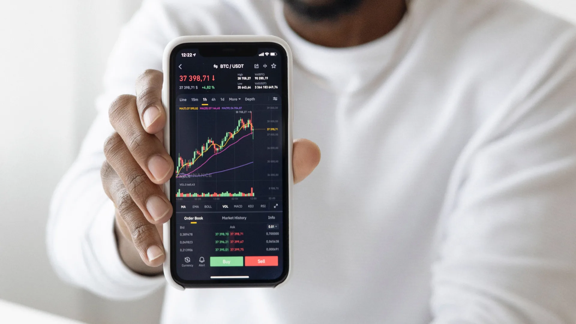 A person in a white shirt holds a smartphone displaying a sleek cryptocurrency trading app, showcasing Bitcoin price movements in true clockwork precision. The screen is filled with green and red indicators, buy/sell buttons, providing an intuitive digital solution for an enhanced customer experience.