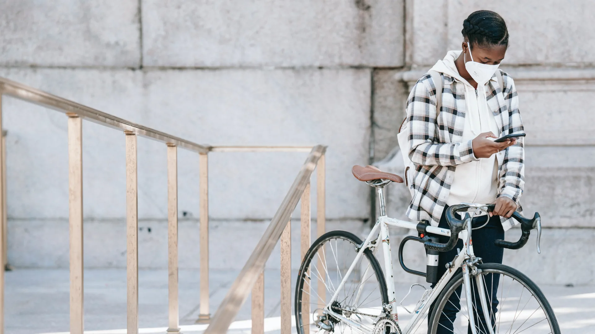 A person in a plaid jacket and face mask stands beside a white bicycle, engaged with their smartphone like clockwork. They are next to a metal railing on a stone-paved surface, with a large stone wall standing impressively in the background.