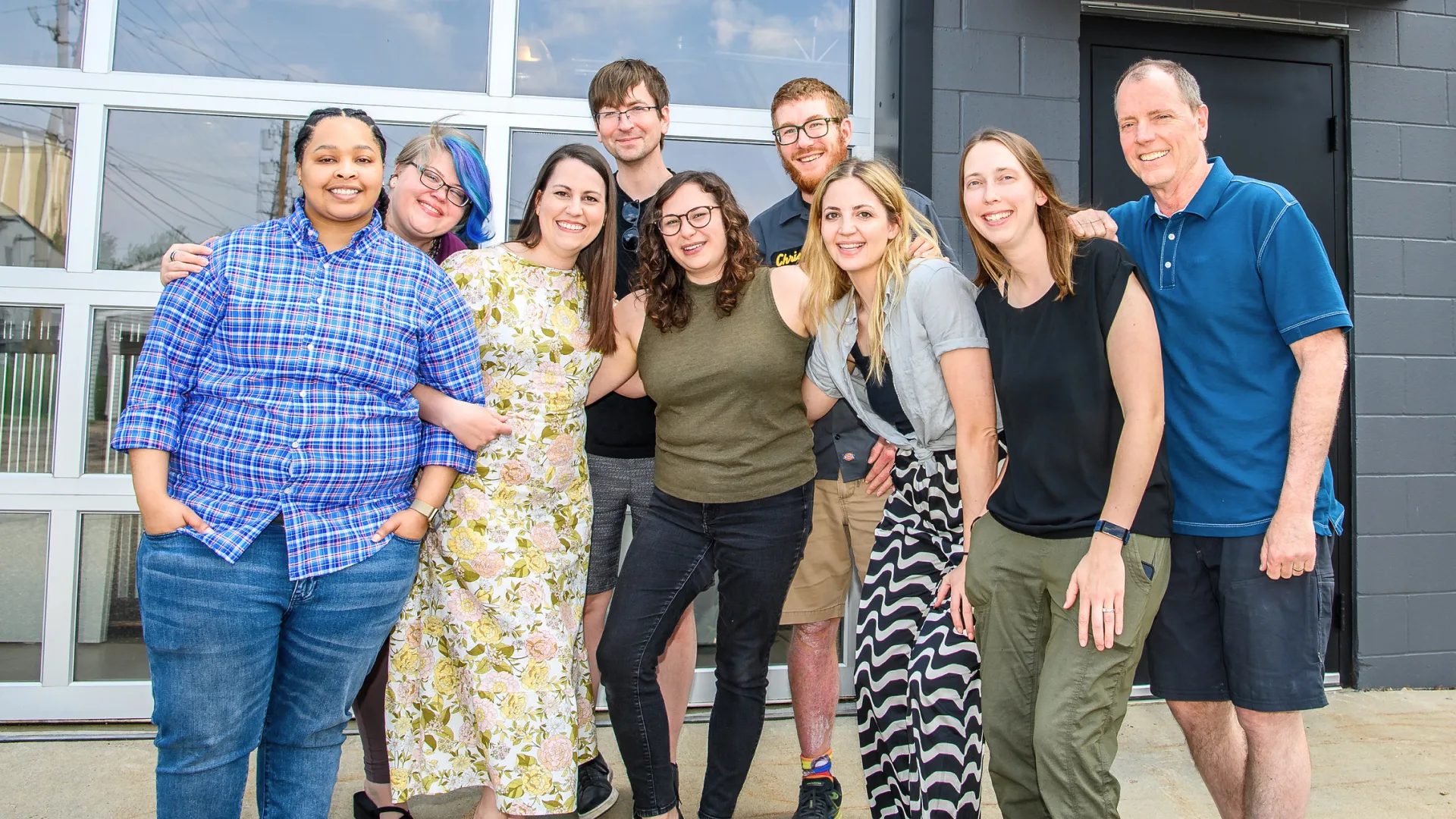 Nine people are standing side by side, smiling at the camera in front of a building with large glass windows. They are wearing casual clothing, and a few have their arms around each other, embodying the spirit of teamwork that fuels innovative experience design.