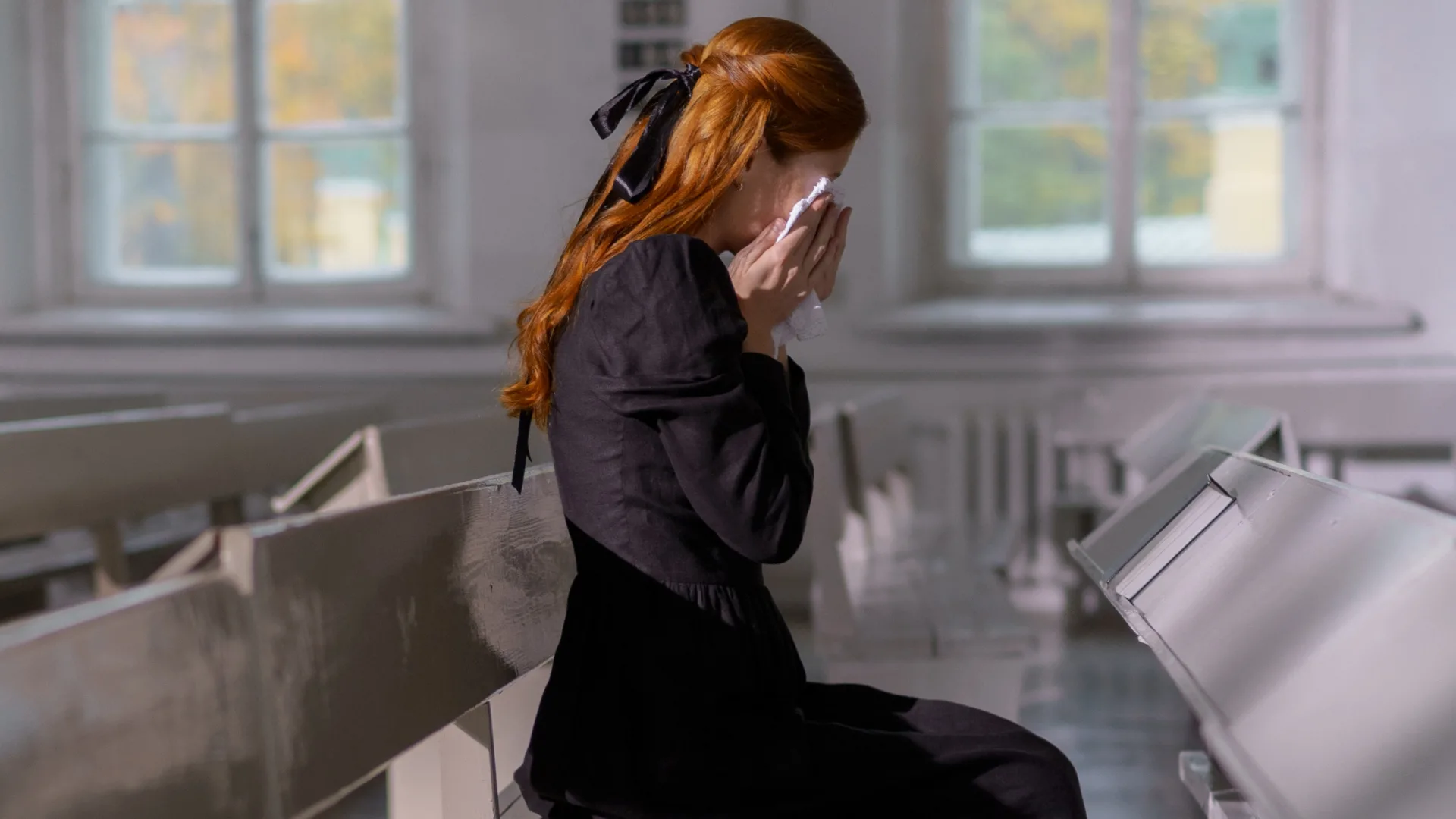 A woman with long red hair, tied with a black ribbon, is sitting on a bench in a bright room. She wears a black dress and uses a white cloth to wipe her eyes, as if shes part of an intricate clockwork of emotions, each tear hinting at the precise movements of sorrow.
