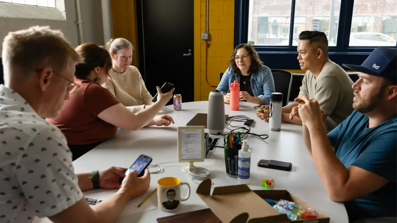 A group of people sits around a conference table covered with assorted items, including drinks and a smartphone. They are engaged in a relaxed conversation, discussing digital solutions, as one person holds a smartphone while another points at something on the table.