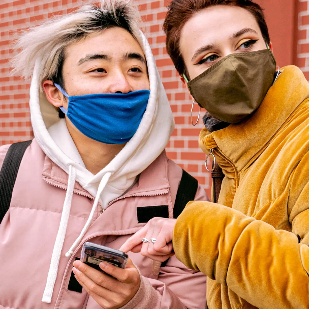 Two people wearing masks are smiling and looking in the same direction. One person with dyed hair is holding a phone and wearing a pink jacket, while the other, in a tan coat, points at it. They stand before a brick building, perhaps discussing the seamless customer experience of custom software.
