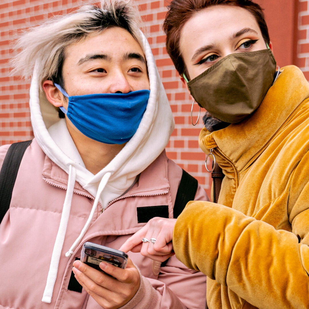 Two people wearing masks are smiling and looking in the same direction. One person with dyed hair is holding a phone and wearing a pink jacket, while the other, in a tan coat, points at it. They stand before a brick building, perhaps discussing the seamless customer experience of custom software.