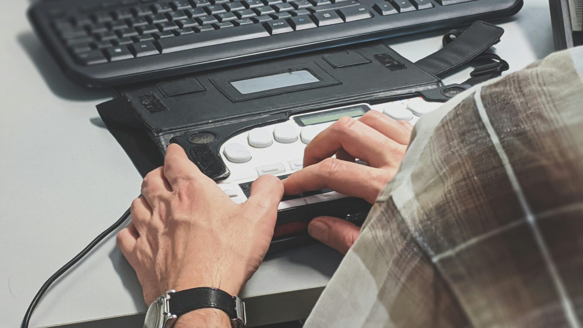 A person uses a braille display and keyboard at a desk, their hands resting on the braille device with a standard keyboard above it, illustrating accessible technology for visually impaired individuals.
