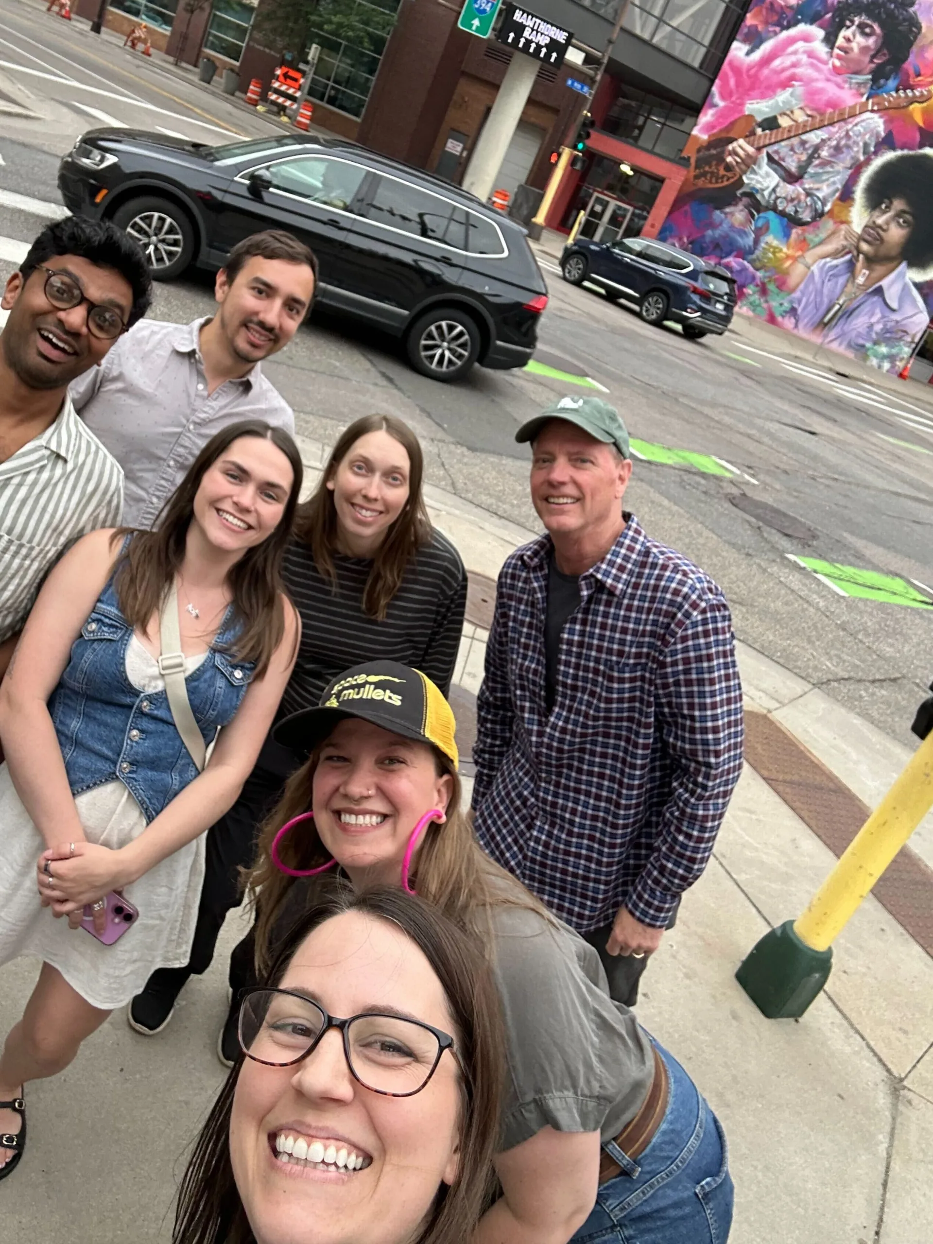 A group of seven smiling people pose for a selfie on a city sidewalk near a crosswalk, with a colorful mural and parked car in the background.