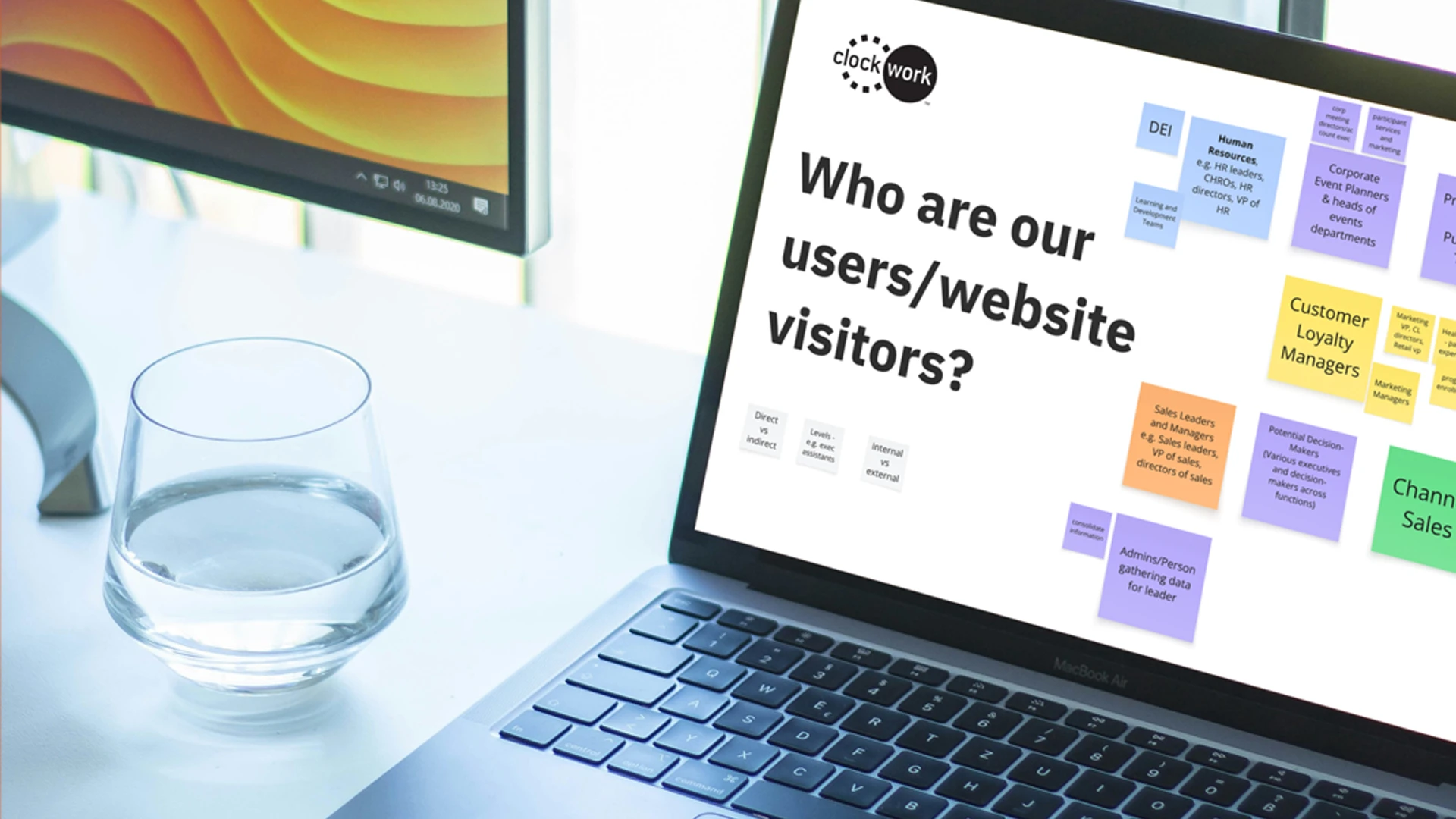 A laptop displays a presentation slide titled “Who are our users/website visitors?” with colorful sticky notes and categories, reflecting insights for a global website. A glass of water and a monitor sit on the white desk beside the laptop.
