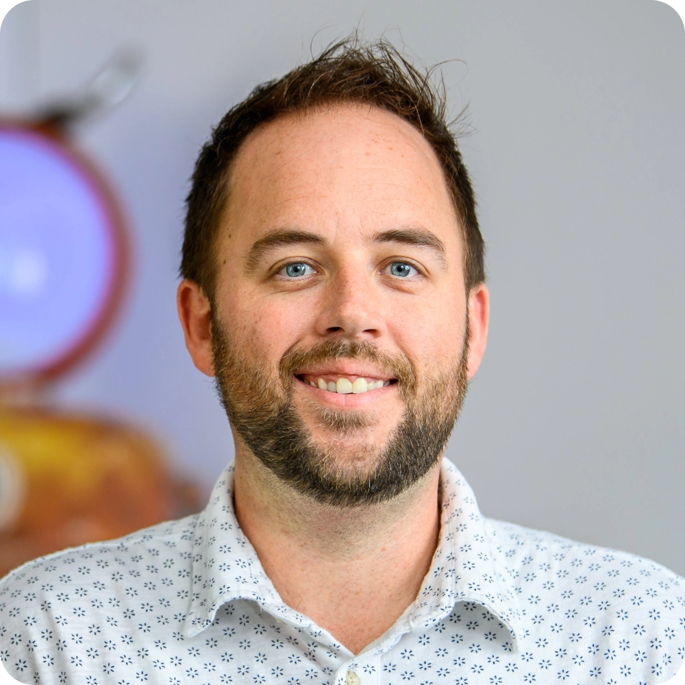 Cassidy Nelemans, Clockwork. A man with short brown hair and a beard, wearing a white shirt with a blue pattern, smiles at the camera against a blurred indoor background—perfectly capturing the approachable style of modern technology consultants.