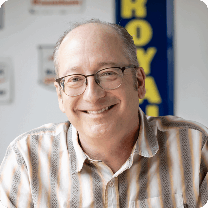 Chuck Hermes, Co-Founder and Chief Experience Officer at Clockwork. A middle-aged man with glasses and a receding hairline smiles at the camera. He is wearing a striped shirt and is seated indoors, with blurred signs—including a blue and yellow ROYAL clockwork sign—in the background.
