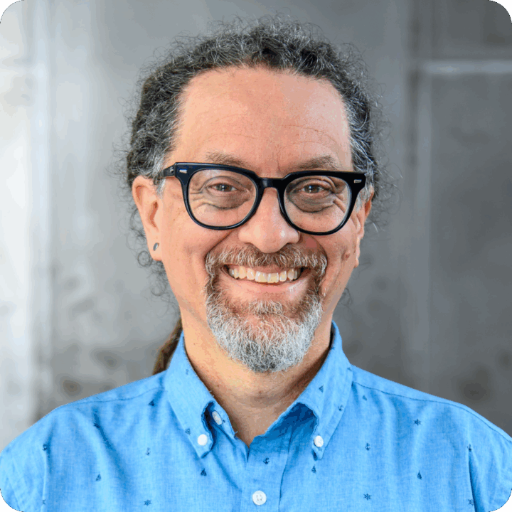Jay Haase, Director of Software Engineering at Clockwork. A smiling person with glasses, a goatee, and curly hair pulled back, wearing a light blue button-up shirt, stands in front of a blurred gray background like clockwork.