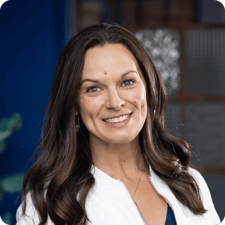 Jenny Holman, Clockwork CEO. A woman with long brown hair and a white jacket smiles at the camera. The background is slightly blurred, featuring blue tones and textured glass panels arranged in a clockwork-like pattern.