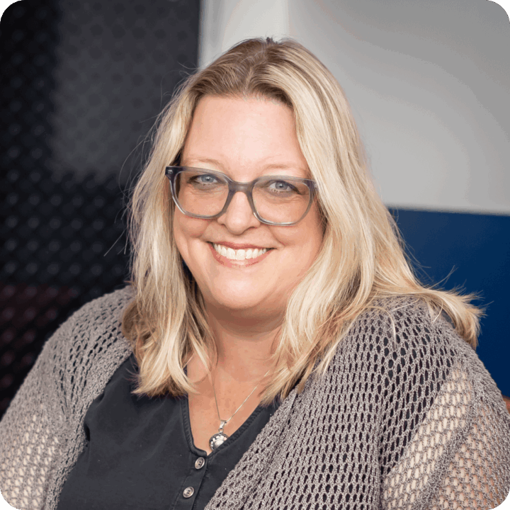 Melanie Shirley, Chief Growth Office at Clockwork. A woman with long blonde hair and glasses smiles at the camera, wearing a black shirt and a knitted gray cardigan, against a blurred, clockwork-inspired background of dark and light colors.