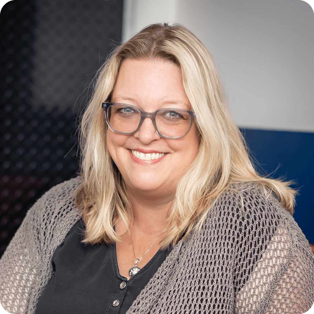 Melanie Shirley, Chief Growth Office at Clockwork. A woman with long blonde hair and glasses smiles at the camera, wearing a black shirt and a knitted gray cardigan, against a blurred, clockwork-inspired background of dark and light colors.