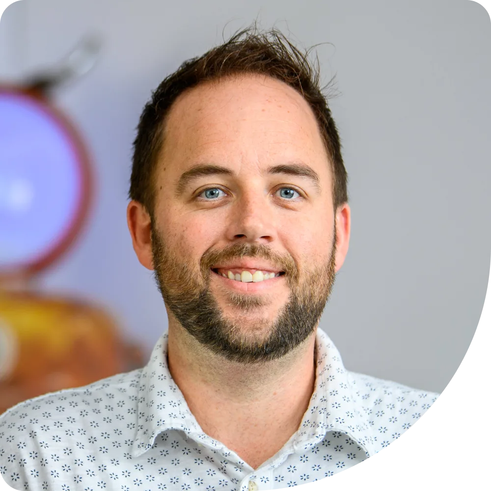 Cassidy Nelemans, Sr. Software Engineer at Clockwork. A smiling man with short brown hair and a trimmed beard wears a white shirt with a blue geometric pattern. The background is softly blurred with hints of purple and brown.