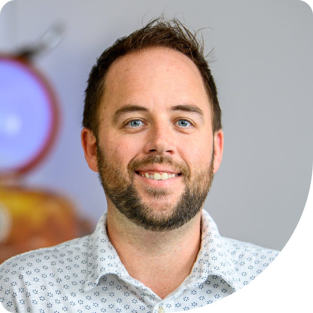 Cassidy Nelemans, Sr. Software Engineer at Clockwork. A smiling man with short brown hair and a trimmed beard wears a white shirt with a blue geometric pattern. The background is softly blurred with hints of purple and brown.