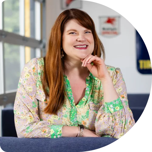 Danielle Miller, Principal User Experience Strategist at Clockwork. A woman with long red hair, wearing a floral blouse with green trim, sits on a blue couch and smiles at the camera with her chin resting on her hand. The background shows a bright, modern indoor space.