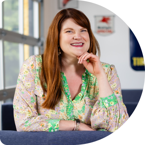 Danielle Miller, Principal User Experience Strategist at Clockwork. A woman with long red hair, wearing a floral blouse with green trim, sits on a blue couch and smiles at the camera with her chin resting on her hand. The background shows a bright, modern indoor space.