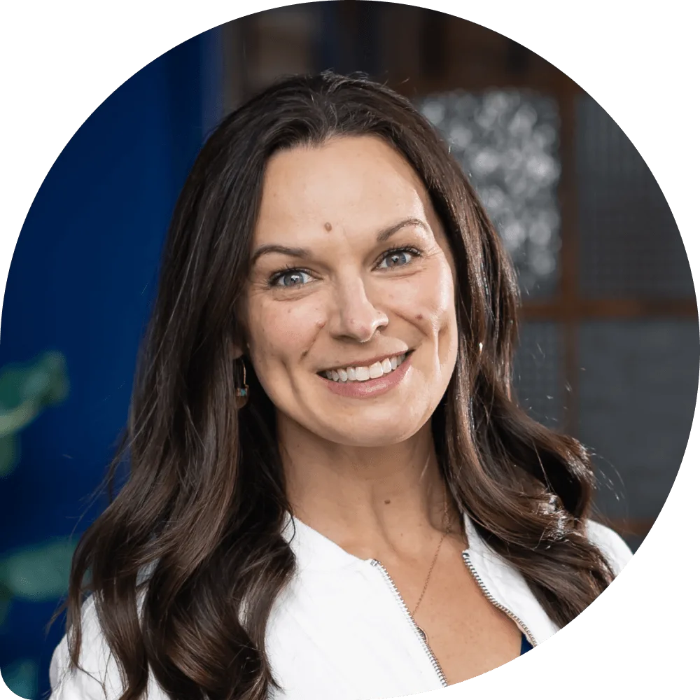 Jenny Holman, Clockwork CEO. A woman with long brown hair and a white top smiles at the camera. The background is softly blurred, showing hints of blue and a window.