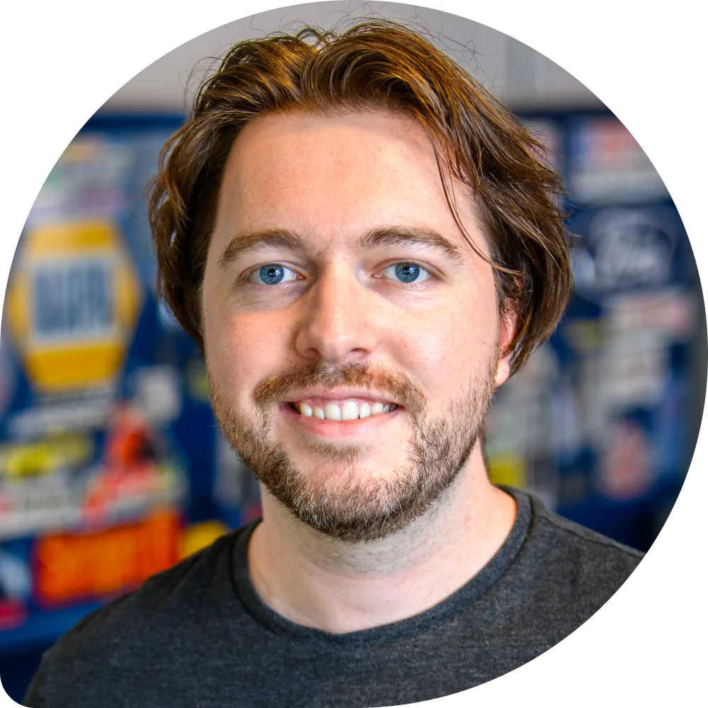 Sam Gordon, Lead Product Designer - Experience and Visual Design at Clockwork. A man with medium-length brown hair and a beard, smiling at the camera. He is wearing a dark t-shirt, and the background is blurred with colorful logos and signs.