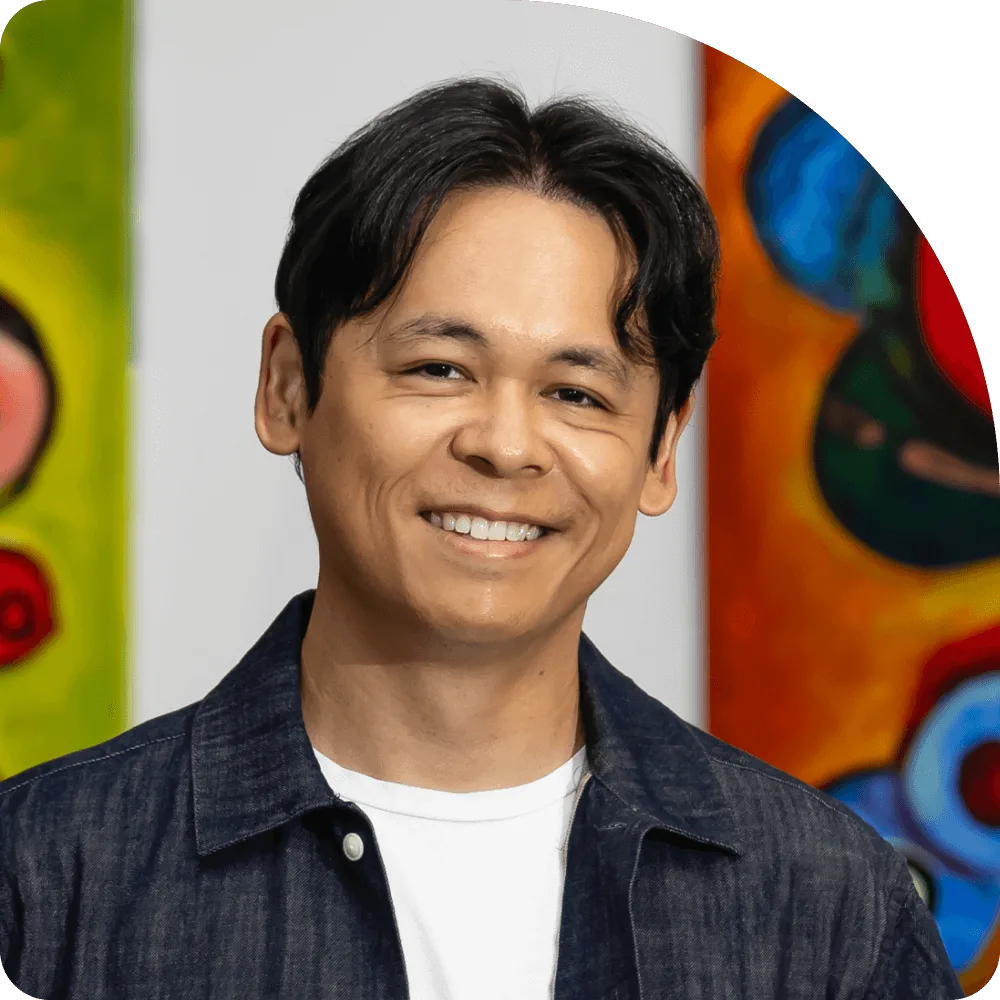 Vince Cabansag, COO at Clockwork. A smiling man with dark hair wearing a white t-shirt and dark jacket stands in front of colorful abstract paintings.