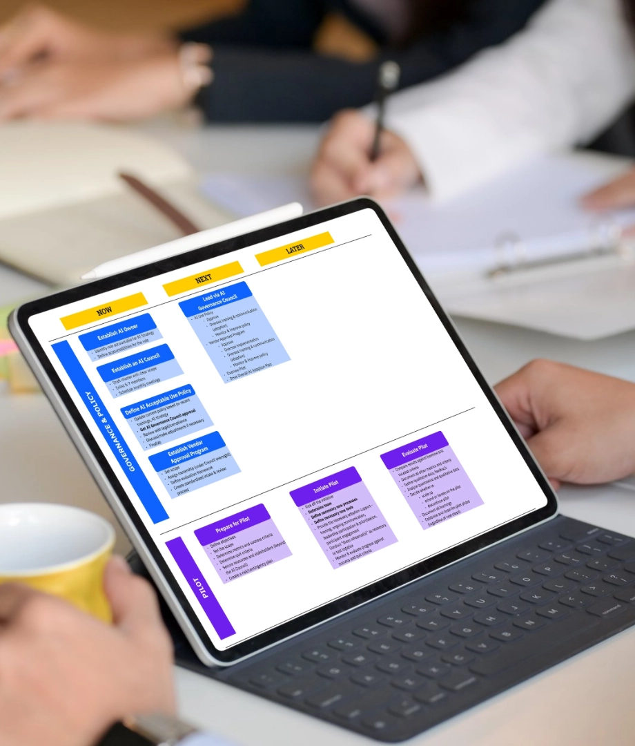 A tablet on a keyboard displays a colorful flowchart as people work at a table with documents, notebooks, and a yellow coffee mug in the background.