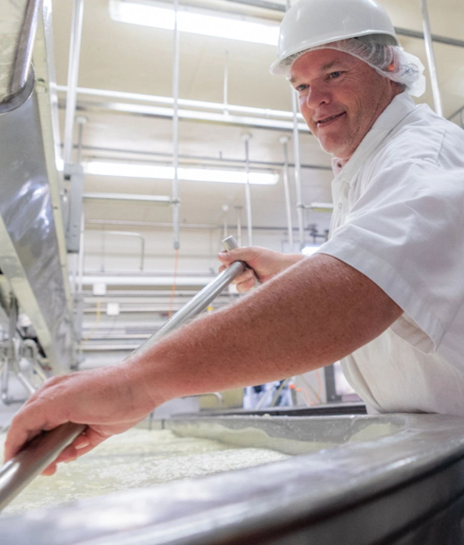 A worker in a white uniform, hairnet, and hard hat stirs a large vat of liquid in an industrial food manufacturer website facility, surrounded by pipes and stainless steel equipment.