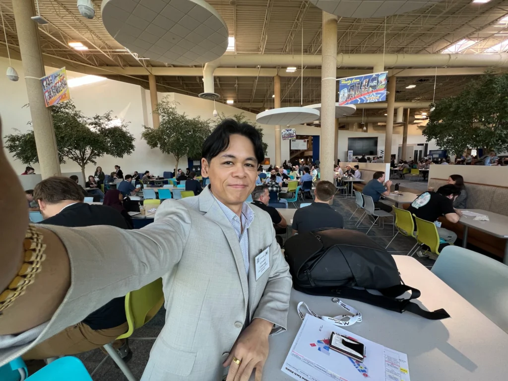 A person in a light gray suit takes a selfie in a busy, modern cafeteria with groups of people at colorful tables. Papers, a phone, and an ai pilot badge are on the table in the foreground.